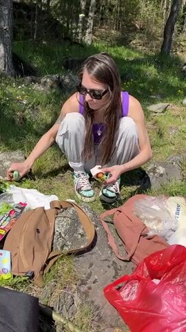 A young woman is depicted squatting on rocky, grassy ground in an outdoor setting, likely preparing for or having a picnic. She wears a purple sleeveless top, light grey cargo pants, and distinctive colorful sneakers with chunky soles. Dark sunglasses complete her casual attire.

The scene is bright and sunny, with dappled sunlight filtering through trees visible in the background, suggesting a daytime event. Around her are several bags: a brown canvas backpack to her left, a reddish-brown tote bag to her right, a white plastic bag with some contents, and a large red plastic bag filled with items.

The woman is actively unwrapping a small, round food item, possibly a falafel or a similar snack, holding it carefully in her hands. She appears focused on the task. At one point, she reaches towards the white plastic bag on her right.

Visible details include:
*   A yellow and blue package near the brown backpack shows "40" and partial text "CEG".
*   The white plastic bag contains some items and partial Cyrillic text "БУМА" (possibly part of "БУМАГА" meaning "paper").
*   A tattoo is visible on the woman's left upper arm.
*   The ground consists of large rocks interspersed with green grass and small yellow flowers.

The overall atmosphere is relaxed, indicating an enjoyable outdoor meal or break in a natural environment near Sliznevo, Russia.