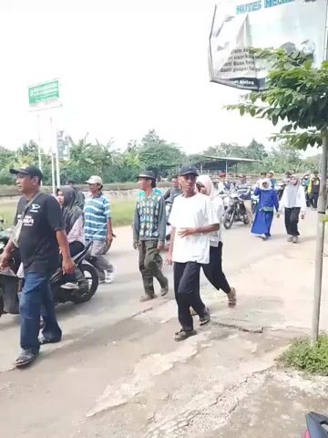The video shows a group of people walking and riding motorcycles down a narrow asphalt road. The majority of individuals are walking. Many of the women in the group are wearing hijabs, and the men are wearing casual clothing like shirts, jeans, and caps. Some people are riding motorcycles, either alone or with a passenger. Two young girls, one wearing a white shirt and blue jeans and the other wearing a white outfit, are walking hand in hand on the side of the road. The environment appears to be a rural or suburban area, with trees and greenery visible in the background. A sign advertising "KUTES HELMET" is prominently displayed on the right side of the road, and another sign is visible in the background. The overall atmosphere appears to be that of a community event or procession.