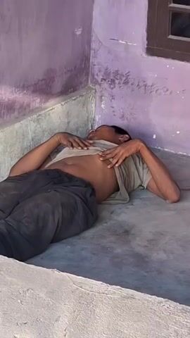 A person is lying on their back on a concrete floor next to a purple wall and a window. They are wearing a beige shirt and dark pants. Their shirt is pulled up, exposing their stomach. Their arms are bent at the elbows and their hands are resting on their chest. The person's head is tilted back, and their eyes are closed. The environment appears to be outdoors, possibly on a porch or patio. The lighting suggests it is daytime.