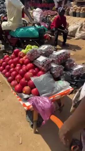 A street vendor displays apples and packaged strawberries on a table with a white and red-striped edge, in what appears to be an outdoor market. A motorcycle with a green cover on the seat is parked next to the table. There is a man in a beige shirt sitting on the motorcycle. Behind the table, another man is sitting on a stool, and in the background are sacks of produce. Other people and produce are visible in the distance, suggesting a busy market atmosphere.