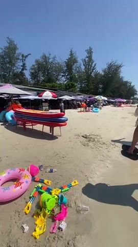 The video begins by showing a beach on a sunny day with a clear blue sky. There are many beach umbrellas set up along the sandy shore, and several people are walking around. In the foreground, there are some inflatable objects, likely for water activities, and various colorful children's toys. As the video progresses, the camera pans to show a man walking along the edge of the water, carrying a pair of flip-flops in his hand. The scene then expands to reveal a crowded beach with many people swimming and playing in the ocean. The water is clear and shallow, with some gentle waves. The overall atmosphere is lively and recreational, indicative of a popular beach destination.