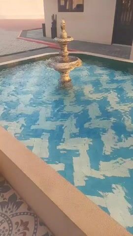 A decorative fountain with tiered basins and a weathered, ornate design stands in the center of a swimming pool. The pool's water is clear and displays a vibrant blue and white geometric tile pattern on its bottom. The fountain is operational, with water gently cascading from the upper basin to the lower one, creating ripples on the water's surface. The surrounding environment suggests an outdoor courtyard or patio area, with cream-colored walls, an arched walkway, a closed dark-colored door, and a decorative lantern hanging overhead. The lighting indicates it's daytime, possibly late afternoon, with warm light casting shadows.