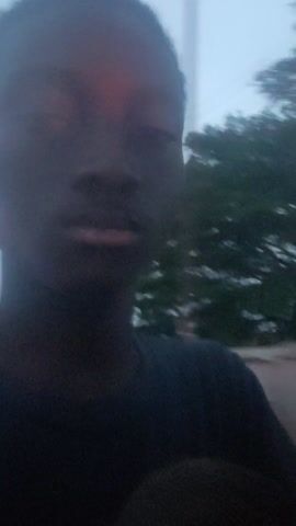 The video shows a person's face, partially visible, against a blurred background of trees and sky. The lighting is poor, and there is a red glow across the face. The person appears to be looking at the camera.