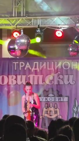 A woman in a sequined dress plays a saxophone on a stage. She is lit by spotlights and surrounded by disco balls. A banner behind her reads "ТРАДИЦИОНОВИЋКИ ЛУКОВИТ" (TRADITIONAL LUKOVIT). The audience is visible in the foreground. The lighting changes colors throughout the video.