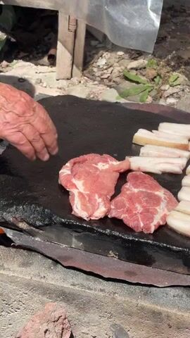 A person's hands place raw meat onto a dark, flat cooking surface. The meat appears to be pork, with both lean cuts and fatty pieces. In the background, a blue bowl holds more raw meat, including what looks like ground meat or sausages. The cooking setup is outdoors, with a makeshift shelter of plastic sheeting and wooden supports visible. The ground is covered in dirt, leaves, and scattered debris. A green plastic bottle is also present under the shelter. The overall scene suggests an informal outdoor cooking or food preparation setting.