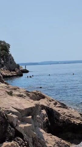 The video shows a rocky coastline with clear blue water and a clear sky. In the distance, there is a hazy shoreline with some trees. Several people are visible in the water, some swimming and others in what appears to be a kayak. One person is standing on a rocky outcrop near the water's edge. The rocks in the foreground are rough and textured, with some appearing to have steps carved into them. The overall scene suggests a sunny day at a natural swimming spot.