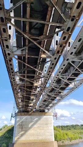 The video shows a low-angle view looking up at the underside of a large metal truss bridge. The bridge spans across a body of water. The sky is blue with some scattered white clouds. The bridge structure is complex, with many interconnected metal beams and supports. The video moves slowly, panning slightly to the right, revealing more of the bridge's length and the surrounding landscape. The landscape on either side of the water appears to be green and wooded. The water itself is a brownish color, suggesting it may be a river. There are no people or vehicles visible on the bridge or in the immediate vicinity. The overall impression is one of industrial scale and engineering.