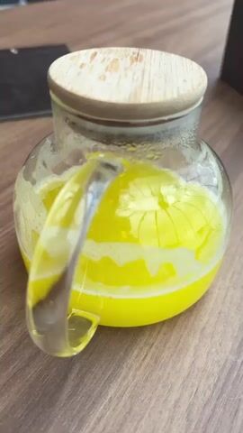 The video shows a wooden table with a glass teapot filled with a bright yellow liquid, likely tea or juice. The teapot has a wooden lid and a clear glass handle. Next to it is a small white plate with a single piece of chocolate on it. A menu and a phone are also on the table. A person places a white cup with a speckled pattern on a matching saucer and fills it with the yellow liquid from the teapot. The person is wearing two silver rings on their left hand. The setting appears to be a cafe or restaurant, with a blurry background showing chairs and a heater.