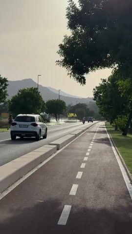 A white car is driving on a road next to a bike path. There are trees on the right side of the road and mountains in the background. A person is running on the left side of the road. The sun is shining and the sky is hazy.