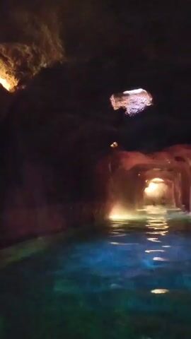 The video shows a water tunnel that appears to be indoors with a dark ceiling and a light illuminating the water and the tunnel ahead. There are people visible in the distance at the end of the tunnel. The water is a blue-green color. The tunnel walls appear to be made of a reddish-brown material. The ceiling has some irregular openings. There are faint sounds of water splashing.