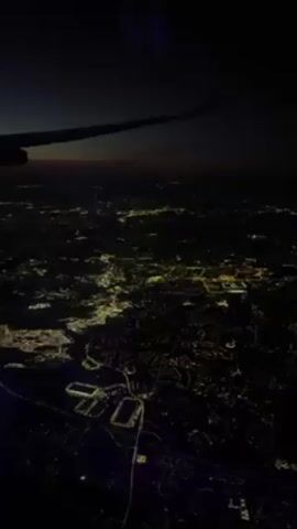 The video shows a night aerial view of a city. The city is brightly lit with numerous lights creating a dense, sparkling network. The camera is positioned inside an airplane, evidenced by the presence of a wing visible in the upper left corner. The night sky is dark, with a faint glow visible near the horizon.