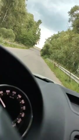 The video is taken from inside a car, showing the driver's perspective. The dashboard, including a speedometer, is visible in the foreground. The car is driving on a paved road that winds through a wooded area with green trees on either side. The sky is overcast.