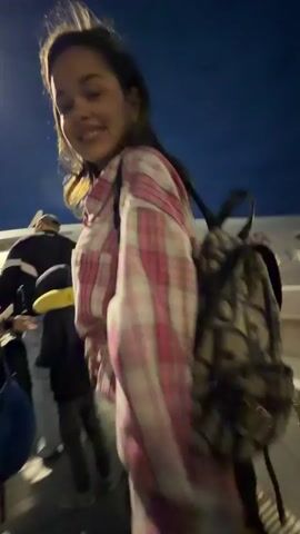 A woman in a pink and white plaid shirt and a man in a beige vest are standing outside at night. They are near an airplane. The woman sticks her tongue out and looks at the man. They then pose together for a video, with the woman kissing the air. Another man wearing glasses is seen standing behind them to the right.