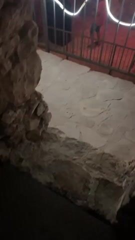 The video captures a dimly lit outdoor area, possibly a patio or courtyard, at night.  The scene is characterized by warm, red lighting that creates a vibrant atmosphere.  The foreground shows a stone-like textured floor and a rough, rocky surface on the left.  A metal railing, decorated with white LED strip lights, runs across the mid-ground.  Beyond the railing, in the background, is a stage or elevated platform.  On this platform, a person, appearing to be a DJ, is standing behind a DJ console.  A large, orange draped fabric is hung in front of the platform, also adorned with strings of small, warm-toned lights.  A disco ball hangs above the DJ, reflecting the ambient light.  The overall impression is that of an outdoor party or event with music and entertainment.  Several indistinct figures are visible behind the railing, suggesting other attendees.