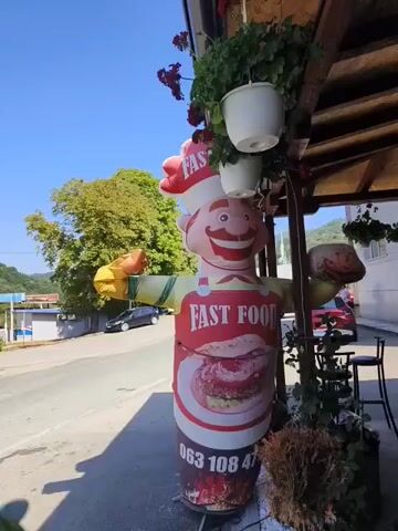 A tall inflatable character resembling a chef stands outside a building, advertising fast food. The character wears a red chef's hat and apron, with a large image of a burger on its torso. The words "FAST FOOD" are printed in red on the apron. A phone number is visible at the bottom of the character's torso. The background shows a street with a car and trees. Potted plants hang from an overhead structure. The sky is blue.
