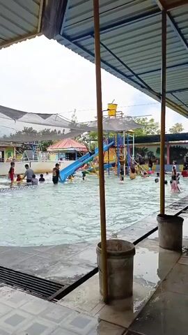 This is a medium-shot, eye-level, static video of an outdoor swimming pool area. A covered shelter with a blue roof and mesh netting extends across the top of the frame. Several people are visible in the pool, some swimming and others wading. There is a blue water slide with yellow accents in the background, and several children are playing on it. The pool area is tiled, and there are two buckets visible in the foreground. The overall lighting suggests an overcast day.