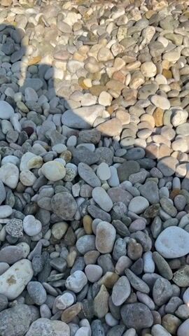 This media file shows a close-up view of a pebble beach with the turquoise sea and a clear blue sky in the background. The pebbles are of various sizes and colors, ranging from white and gray to brown and pink. The sun is shining, casting shadows on the pebbles, indicating it is likely daytime. In the distance, faint outlines of mountains can be seen. A person is visible in the water, enjoying the sea, with a small wave breaking near them. The overall scene conveys a sense of a peaceful and beautiful coastal environment, typical of a summer day at the beach. There is no visible text in the media file. The location context is Općina Baška, Croatia.