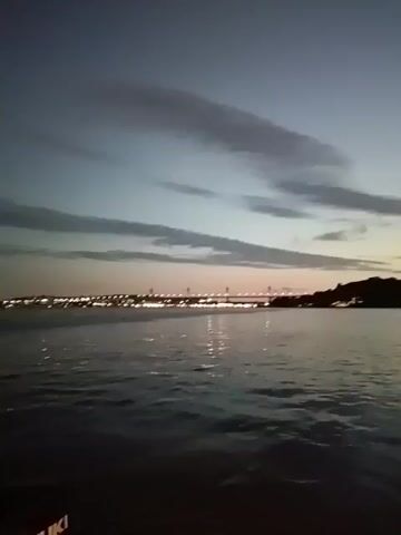 The video shows a body of water, possibly a lake or a bay, under a sky at dusk with long, streaky clouds. In the distance, a bridge is visible, adorned with lights. The lights are reflected in the water, creating a shimmering effect. The camera appears to be on a boat, as the Suzuki logo is visible in the bottom left corner of the frame. The camera is shaking slightly, indicating movement. There are some faint voices in the background.