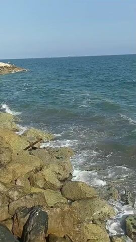 A video shows a rocky shoreline with waves crashing. In the background, there is a clear blue sky and a pier with white umbrellas. There are a few people sitting on the pier and on the rocks. The water is a vibrant blue and the rocks are a mixture of gray and yellow. The scene is peaceful and serene.