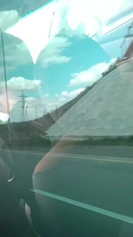 This video is an interior shot from a vehicle looking out the window. The window reflects the interior of the vehicle, with what appears to be a seat or seats and parts of the ceiling visible. Outside the window, there is a landscape with a road, power lines, and vegetation. The sky is blue with white clouds. The camera appears to be moving, as evidenced by the slight blur of the outside scenery.