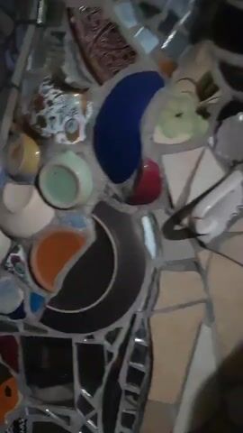 The video shows a mosaic made of broken ceramic pieces, including cups, plates, and bowls, as well as mirrored tiles. The mosaic appears to be on a vertical surface, possibly a wall or a sculpture. The colors are varied, with blues, greens, yellows, oranges, whites, and browns being prominent. Some of the ceramic pieces have floral patterns or other decorative designs. The lighting is dim, and the camera is moving, creating a slightly blurred effect.