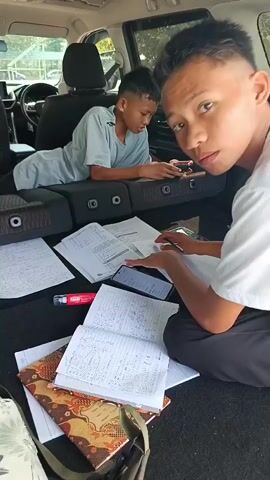 The video shows two young males in the back of a vehicle, surrounded by papers that appear to be schoolwork or notes. The vehicle's rear seats are folded down, creating a flat surface. One male, wearing a light gray t-shirt, is lying on his side on the folded seats, looking at a smartphone he is holding. Papers are scattered around him and on the seats. The other male, wearing a white t-shirt and dark pants, is sitting cross-legged on the floor of the vehicle, with papers in front of him. He is holding a pen and appears to be writing or reviewing the papers. Another smartphone is visible on the floor between the two individuals. The environment outside the vehicle suggests it is daytime, with trees and sunlight visible through the windows. There is a sense of an impromptu study session or homework completion taking place within the vehicle.