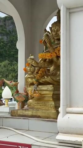 A close-up, eye-level shot features a golden statue of a multi-armed deity seated on a tiered pedestal. The statue is adorned with garlands of orange and white flowers. In front of the statue, to the left, are two decorative, colorful mythical bird-like figures with open beaks and crests. The statue and figures are housed within an open-air structure with white, curved pillars and arched openings. Through the openings, a lush green, tree-covered mountainside is visible under an overcast sky. In the background, a white vehicle is partially visible. The foreground shows white tiled flooring and a white cord or rope.