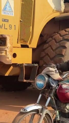 The video shows a large yellow front-end loader with a man walking beside its massive tire. The camera then pans to show the interior of the loader's cabin and the operator. Next, a small motorized tricycle is seen carrying concrete blocks, with the loader in the background. The scene then shifts to show the front-end loader in the process of dumping sand into the back of a large orange dump truck, while a person on a motorcycle observes. Finally, the dump truck is shown driving away with its bed raised. The environment appears to be a construction site or an area with unpaved roads and construction materials.