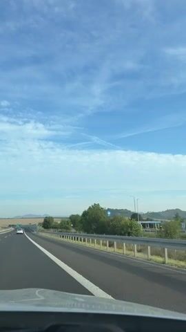 The video shows a first-person perspective of driving on a highway. The sky is blue with scattered white clouds. The road is asphalt with white lane markings. There are guardrails on the right side of the road, and some trees and vegetation beyond them. There are a few cars visible on the highway ahead. In the distance, there are rolling hills and fields. The video captures the movement of the car as it travels down the highway.