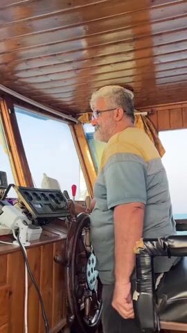 A man with graying hair and a beard, wearing glasses and a t-shirt, stands at the helm of a boat, his hands on the ship's wheel. He is in the captain's cabin, which features wooden paneling on the ceiling and walls, and large windows. To his left are various navigational instruments. The man turns his head to look at the camera and then resumes his focus on steering.