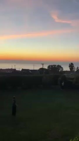 The video shows two people playing catch in a backyard at sunset. The backyard has a green lawn and is bordered by a hedge. Beyond the hedge, there are houses, trees, and a view of the ocean. The sky is a mix of blue, orange, and pink. One person is standing near the left side of the frame, and the other is near the right side. The person on the right throws a ball toward the person on the left, who catches it.