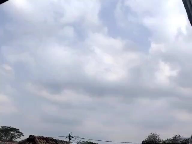 The video shows a partly cloudy sky with the sun intermittently visible through gaps in the clouds. The lower part of the frame displays the tops of buildings, trees, and power lines. The overall scene is outdoors, suggesting it's daytime.