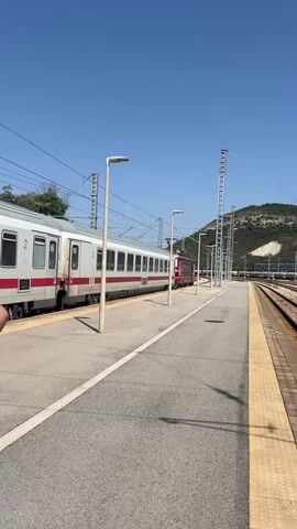 A train with white and red cars is stopped at a train station. The train is on the left side of the frame, and the platform is in the center. There are several lamp posts on the platform. To the right of the platform are multiple train tracks. In the background, there is a hill with some trees and a clear blue sky. The train is stationary.