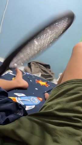 The video shows a close-up view of a tennis racket, held by a person whose arm and leg are visible. The racket is black and has a white mesh. The background is a blue wall and green fabric. The scene appears to be indoors.