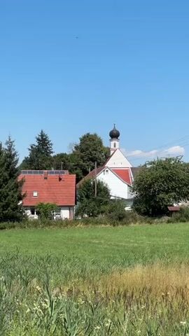 The video shows a rural landscape on a clear, sunny day. In the foreground, there is tall grass and a dirt path. In the midground, there is a red-roofed building with solar panels, and behind it, a white church with a black dome and red trim. Trees surround the buildings. The background is a clear blue sky. The camera pans slightly to the right, revealing more of the field.