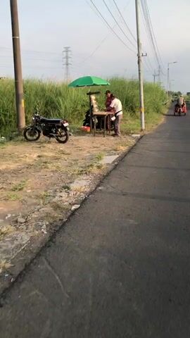 The video shows two individuals standing at a roadside stand displaying various items, possibly cigarettes. A green umbrella provides shade over the stand, which is a wooden table with a tiered display rack. One person, wearing a white t-shirt and a cap, is seen interacting with the items on the display. Another person, also wearing a cap, stands behind them. A motorcycle is parked to the left of the stand, and tall grass lines the background. The scene appears to be outdoors, near a road, during daylight.