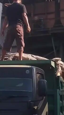 A man wearing a black t-shirt, brown shorts, and a cap stands on top of a load of goods on the back of a green truck. He has a cigarette in his mouth and is holding a red and white flag. The truck is parked in front of a corrugated metal building with some machinery visible in the background. The man walks across the load of goods and then crouches down.