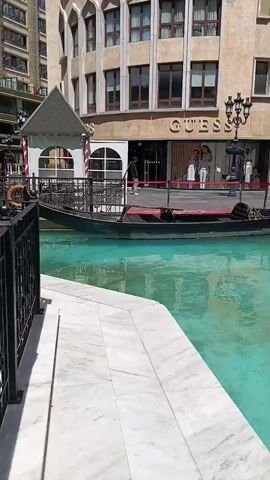 A long, dark gondola boat is partially visible in a narrow, turquoise canal. The canal is bordered by a white marble walkway on the left. In the background, a multi-story building with a light brown facade and arched windows stands. A store with a large "GUESS" logo is visible on the ground floor. A single street lamp is positioned between the canal and the store. Two figures are walking on the sidewalk in front of the store. The scene appears to be an outdoor setting on a sunny day.
