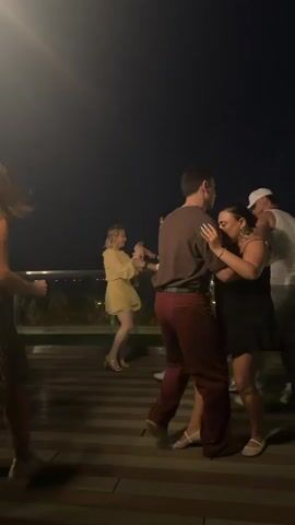 A group of people are dancing on an outdoor patio at night. The patio has a wooden floor. In the foreground, a man wearing a brown shirt and maroon pants dances with a woman in a black dress. Behind them, a couple dances, and further back, another couple dances. In the background, the lights of a city are visible. The sky is dark.