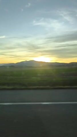 The video shows a view from a moving vehicle on a highway, facing a flat, green field, with a mountain range on the horizon. The sun is setting behind the mountains, casting a golden glow. The sky is partly cloudy, with blue and gray tones. A metal guardrail is visible along the roadside.