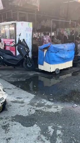 The video shows an outdoor market scene. A black scooter is parked next to a juice stand. A man on a motorcycle is seen approaching from the left. In the foreground, a person is lying on the ground next to a curb. The market appears to be set up with stalls and vendors. The ground is wet and shows puddles, suggesting recent rain or cleaning. The overall atmosphere is that of a busy marketplace.