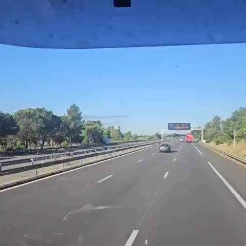 The video shows a view from a car traveling on a highway.  There are green trees and a metal guardrail along the side of the road.  A car is driving in the right lane.  In the distance, smoke can be seen in the sky, with mountains behind it. As the camera angle shifts upward, two planes can be seen flying in formation in the distance.  A utility pole with wires is in the foreground.