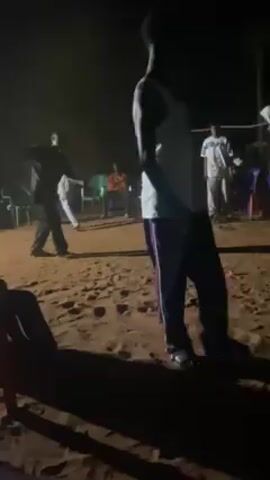 The video captures a night scene outdoors, where a group of people has gathered on what appears to be sandy ground. Several individuals are visible, with some seated in chairs around the periphery. In the center of the frame, two people are engaged in dancing or some form of physical activity under dim lighting. One person, seen closer to the camera, is wearing a white tank top and dark trousers, while another person is in darker clothing, making it difficult to discern specific details. There are some green and other colored chairs around the ground where they are gathering. Overall, the scene conveys a sense of a casual outdoor event or gathering in a low-lit environment.