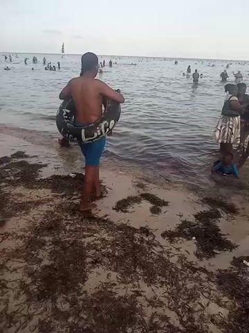 The video shows a beach scene where several people are wading in the shallow water, with many more visible further out. Two men, shirtless, are walking into the water carrying inflatable swim rings with patterned designs. A woman in a white shirt and a girl in a skirt are seen on the left of the men, while a group, including a child, is gathered to the right. There appears to be seaweed scattered along the shoreline. In the distance, one can spot a sailboat, and the sky is overcast.