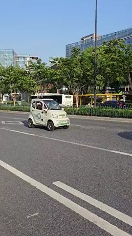 A white electric car is traveling on a road. The car has green license plates and a sticker on the door. There are other cars and a bus in the background, as well as trees and buildings. The sky is blue.