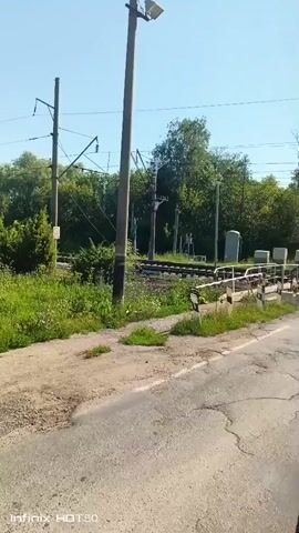 The video captures a bright, sunny day with clear blue skies. The scene is dominated by a rural or suburban landscape featuring a set of train tracks that disappear into lush green foliage and trees. Overhead power lines for the electric train are visible, supported by tall utility poles. A prominent, thick utility pole stands in the foreground, slightly off-center to the left. To the right of the train tracks, a white and grey metal structure, possibly a barrier or part of a crossing, is present. The ground in the foreground is unpaved and dusty, with patches of green grass growing through. A cracked asphalt road runs along the bottom of the frame. In the middle ground, a red and grey train locomotive is moving along the tracks, partially obscured by trees. The train appears to be an electric locomotive due to the overhead power lines. The overall impression is one of an outdoor scene by a railway line on a pleasant day.