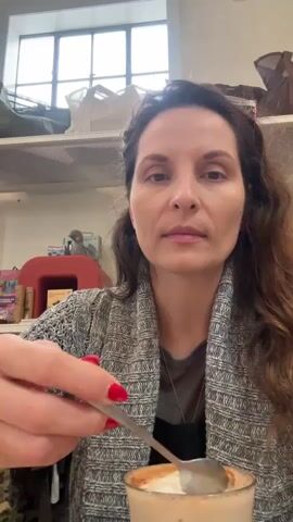A woman with dark wavy hair and red nail polish holds a spoon with a dollop of whipped cream and a small piece of pink candy. She brings the spoon to her mouth and eats the contents. She is wearing a grey knit sweater over a darker shirt. In the background, a glass of a frothy beverage is visible, along with shelves filled with items. The environment appears to be indoors, possibly a cafe or shop.