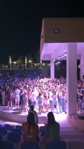 This is a vertical, nighttime video taken from behind a crowd of people, looking towards an outdoor stage area. A large group of mostly young people, dressed in casual summer clothing, are gathered on and around a raised stage. The stage is lit with pink and white lights, and there are rows of blue stadium-style seats in the foreground and background, with some people sitting in them and others standing. The people on the stage and in the surrounding area appear to be dancing and interacting. The overall atmosphere seems to be that of a concert or outdoor party. The video is short and captures a dynamic scene.