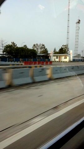 The video shows a view from inside a vehicle, likely a car or bus, looking out a window. The perspective is slightly tilted, and motion blur is evident, indicating the vehicle is in motion. The scene outside includes a paved road with white markings, a barrier system made of grey concrete blocks with colored accents, and behind that, some buildings and trees. There are also tall structures resembling towers or poles in the distance. The lighting suggests it is daytime, possibly overcast or at dusk/dawn. There are no visible people or animals. The glass of the window reflects the interior of the vehicle, including a curved edge at the bottom left, which might be part of the window frame or the dashboard.
