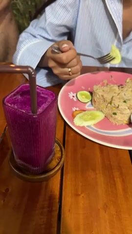 The media file is a close-up shot of a table in what appears to be a restaurant in Tong Yang, Thailand. On the wooden table, there is a glass of vibrant purple smoothie with a brown straw. Next to the smoothie is a pink plate with a floral design, containing a portion of fried rice, slices of cucumber, and lime wedges. A person, wearing a blue and white striped shirt, is visible in the upper right portion of the frame. Their hands are engaged in holding a fork, which is positioned above the plate, suggesting they are in the process of eating or about to eat the fried rice. The lighting suggests it might be evening or indoors, as the light is soft. The overall mood appears relaxed and casual, indicative of a meal being enjoyed. No visible text is present in the media file.